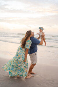 Baby being lifted in air by dad as mom watches on the beach
